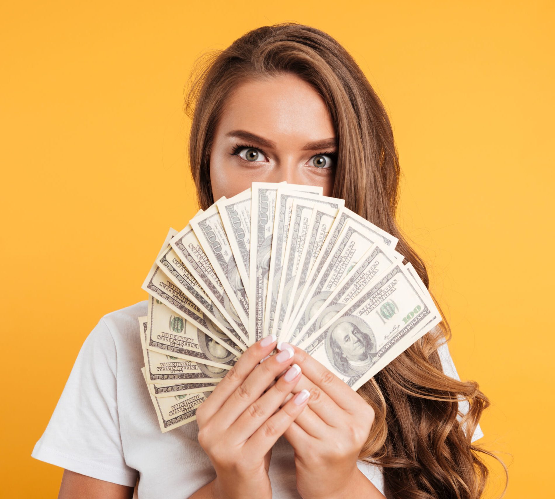 Close up portrait of a young girl covering her face with money banknotes isolated over yellow background